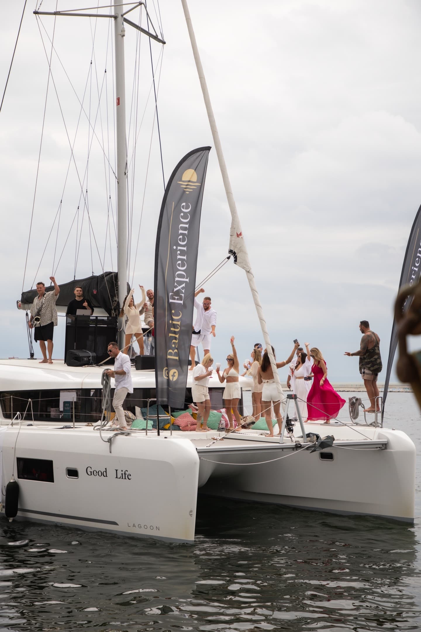 Szerokie ujęcie imprezy na katamaranie Good Life z flagą Baltic Experience, tańczący goście w bieli i kobieta w czerwonej sukience, DJ i saksofonista na pokładzie.